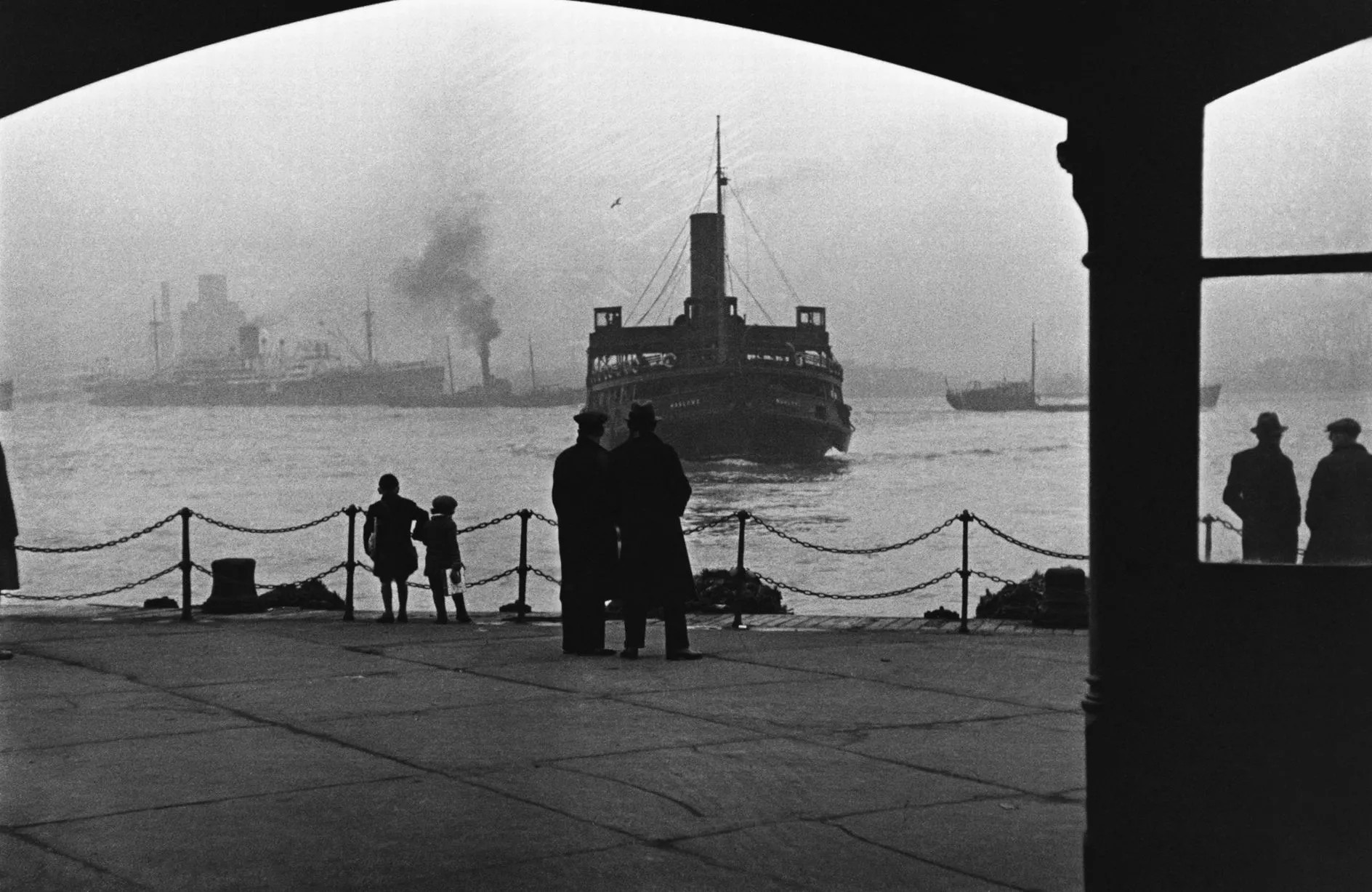 A black and white photo from the 1930s of a ferry approaching a pier, with silhouettes of two adults and two children waiting and watching.